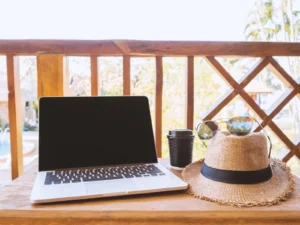 A Laptop, sunglasses, coffee, and a hat sitting on a table outlooking a patio