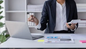 A woman standing at a laptop pointing towards the screen with a notepad in the other hand